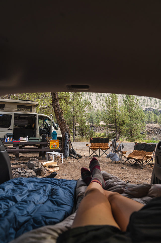 View from the back of a vehicle showing a person's legs in a sleeping bag, looking out at a campsite. The campsite includes a van, folding chairs, a table with supplies, and a cooler. The surrounding area has trees and a mountainous landscape.