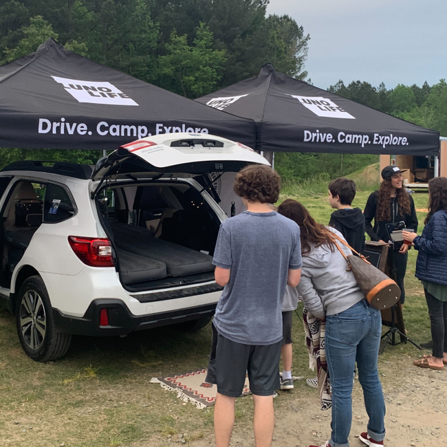 People gathered around the back of a white SUV with an open trunk, under two black tents that read "Drive. Camp. Explore." in a wooded outdoor setting. A man in a blue shirt and shorts is at the center, with others observing and interacting.