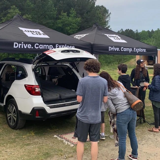 A group of people stands around the open trunk of a white SUV, parked under two black tents with the logo "Luno Life" and the slogan "Drive. Camp. Explore." in a grassy outdoor area with forest in the background.