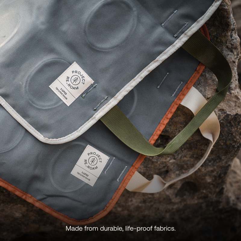 Two durable Firewood Carrier Utility Mats from Luno HQ are lying on a rock. These eco-friendly accessories feature tags that read "PROJECT ROAM" and "Eco Living." One mat has a white trim, and the other has a brown trim. Text at the bottom says, "Made from durable, life-proof fabrics.