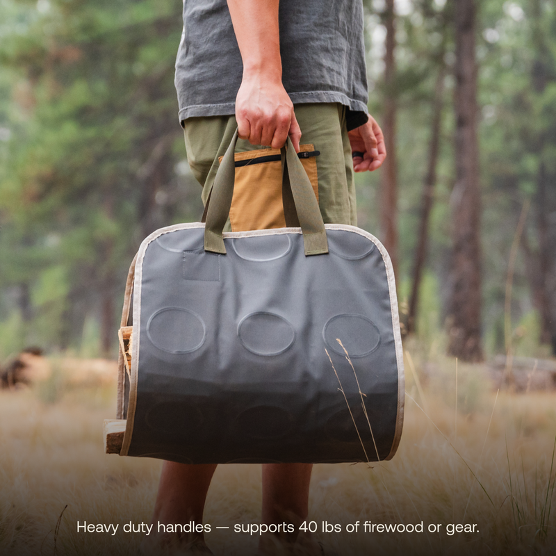 A person uses heavy-duty handles to carry a Firewood Carrier Utility Mat by Luno HQ, made of durable fabric and designed to support up to 40 lbs of firewood or gear. The person is standing in an outdoor setting with trees and grass in the background. The text at the bottom reads, "Heavy duty handles — supports 40 lbs of firewood or gear.