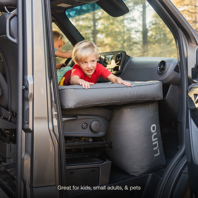 Two young children play on a makeshift bed inside the rear of an SUV. One child in a red shirt lies on the elevated platform, while the other is visible in the background. The setup, featuring Luno's® AIR Van Front Cab Mattress with durable fabric, is great for kids, small adults, and pets.