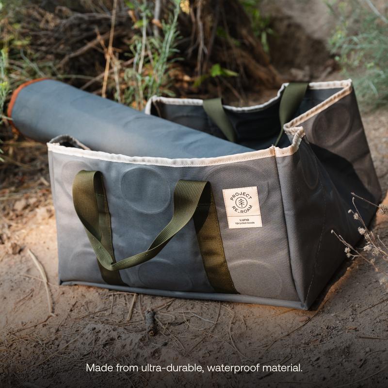 Two brown and green waterproof bags with a durable structure are placed on the ground among dried leaves and shrubs. One bag, a 50L Gear Tote from Luno HQ, contains a rolled gray mat. A label on one bag reads "Wander Pro Kit - Project Water." The alt text at the bottom states the material's durability.