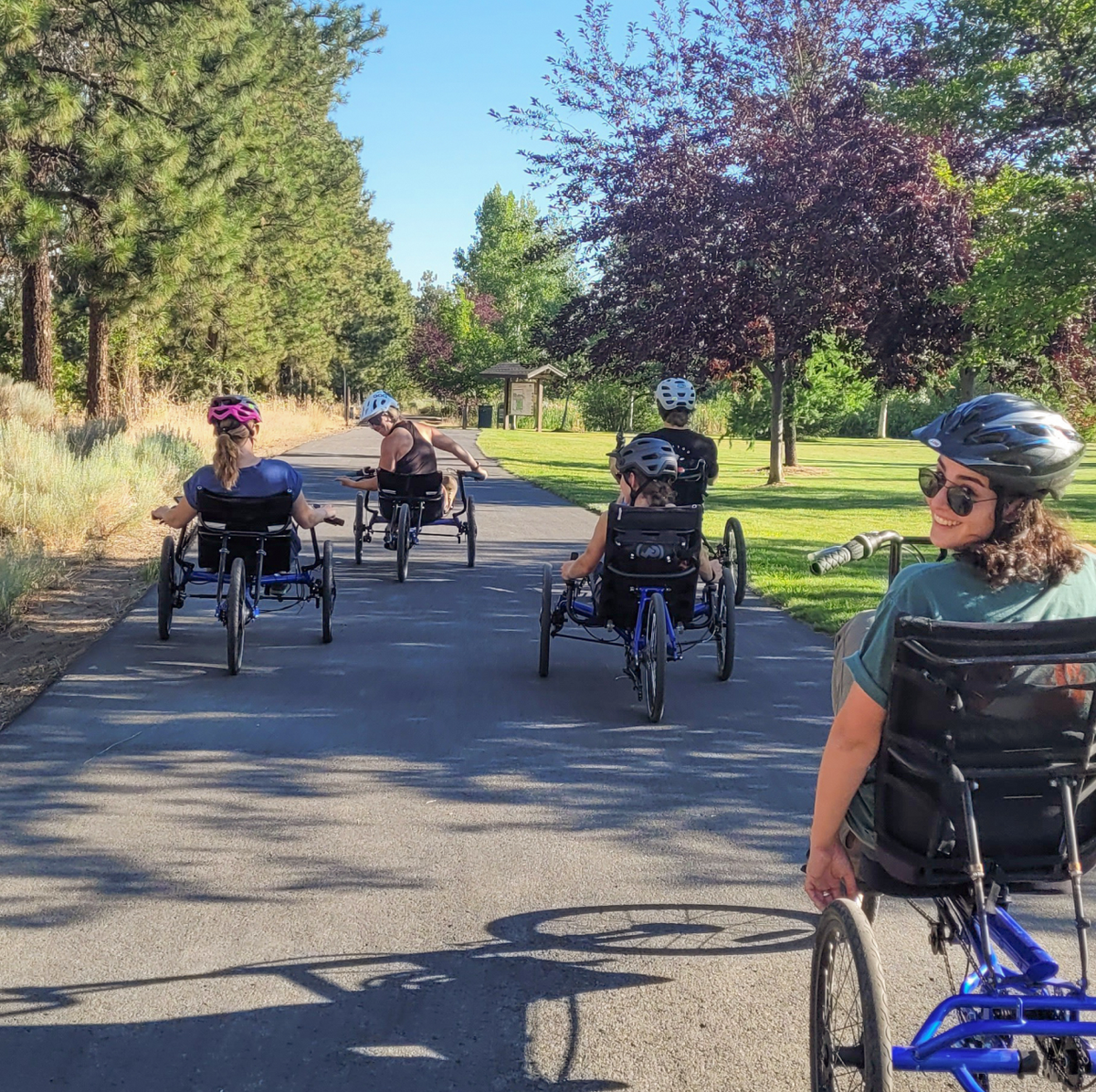 A group of people ride adaptive hand cycles on a paved path surrounded by trees and a grassy area on a sunny day. The cyclists are wearing helmets, and one of them is glancing back and smiling at the camera.