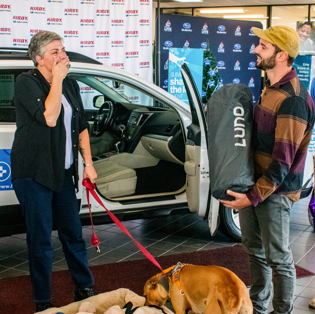 A woman covering her mouth in surprise holds a dog's leash while standing beside a car with its door open. A smiling man wearing a striped sweater and cap holds a packaged item labeled "LUNO" in front of a backdrop with logos. A dog lies on the floor.