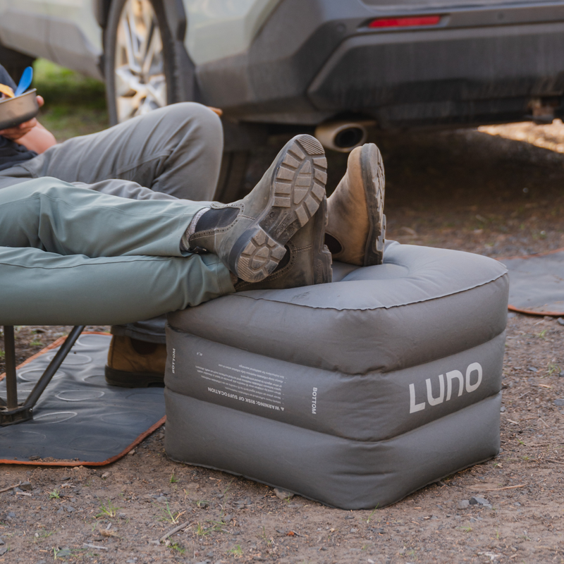 Two people relaxing outdoors with their legs propped up on gray Head Support Space Filler Cubes (Pre-Owned) branded with "Luno HQ." They are sitting next to a parked vehicle, resting on outdoor mats that could double as a comfortable sleeping surface. The ground is covered with dirt and small rocks.