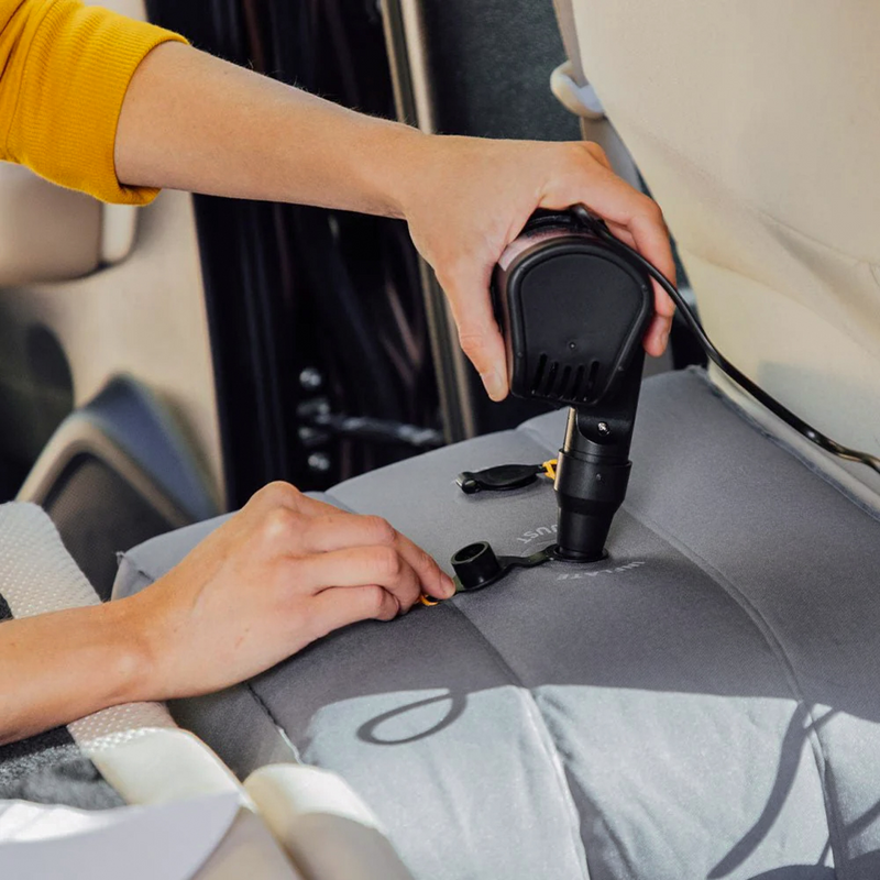 A person inflates a set of Head Support Space Filler Cubes (Pre-Owned) by Luno HQ on a car seat using an electric air pump. The black air pump is connected to the car via a cord. The person's hands adjust the valve on the cubes, ensuring they are properly inflated. The car's interior is visible in the background.