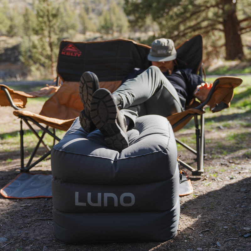 A person relaxes outdoors on a black inflatable ottoman with the brand name "Luno HQ" visible. Their feet are propped up, they're wearing a hat and casual clothing, and they are seated in a foldable camping chair beside Head Support Space Filler Cubes (Pre-Owned), with a natural, sunny backdrop of trees and grass.