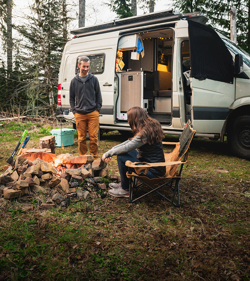 A person stands next to an open camper van while another person sits by a campfire on a folding chair. The surrounding area is a forest with green trees and a patch of grass. Firewood is stacked near the campfire. Camping equipment is visible near the van.