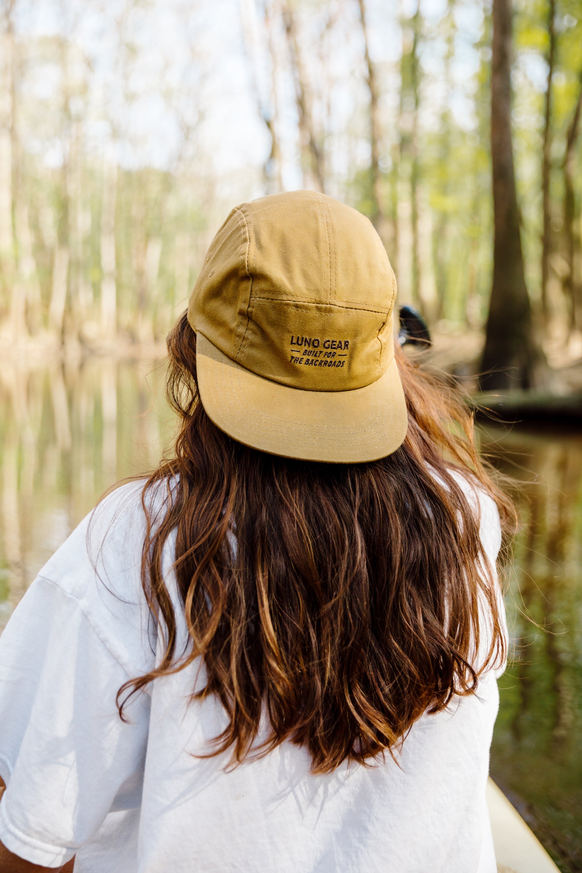 A person with long brown hair, wearing a beige cap with "Luno Gear" written on it, is seen from behind. They are sitting in a boat surrounded by a wooded area with trees reflecting off the water. The person is also wearing a white shirt.