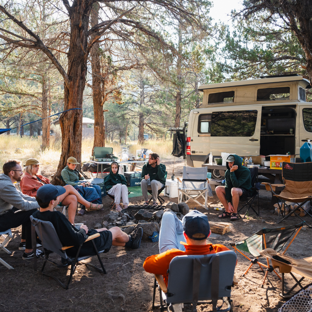 A group of people sit around a campfire in a wooded area with tall trees. They are camping near a white camper van. Chairs, camping equipment, and coolers are scattered around, and some people wear hats and jackets, suggesting a cool day.