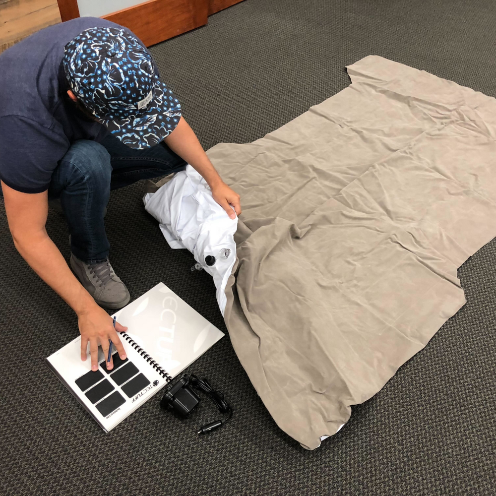 A person in a blue patterned cap and jeans, kneeling on a carpeted floor, examines fabric laid out in front of them. They are holding a fabric sheet with one hand and looking at a folder with fabric swatches. A black camera and camera strap lie nearby.