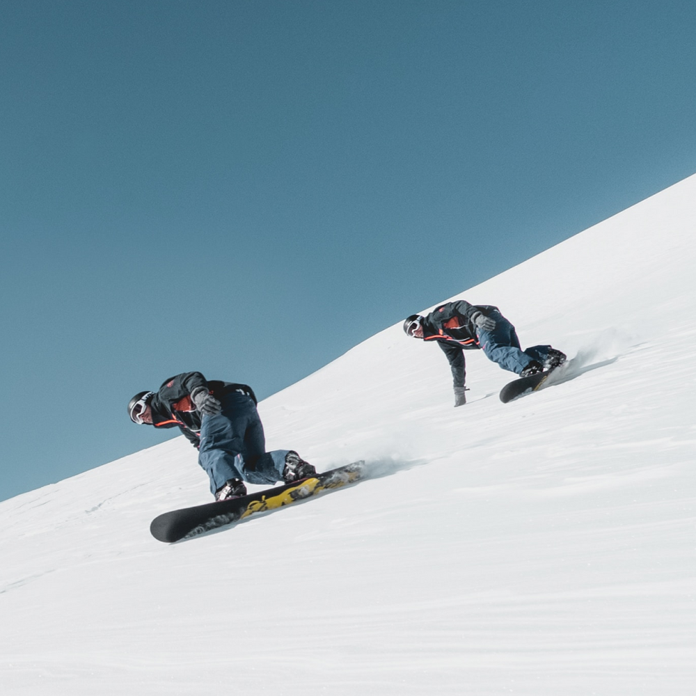 Two snowboarders descend a snowy slope against a clear blue sky. Both wear helmets and winter gear, carving through the snow at an angle. The image captures motion and the expanse of the snowy terrain.