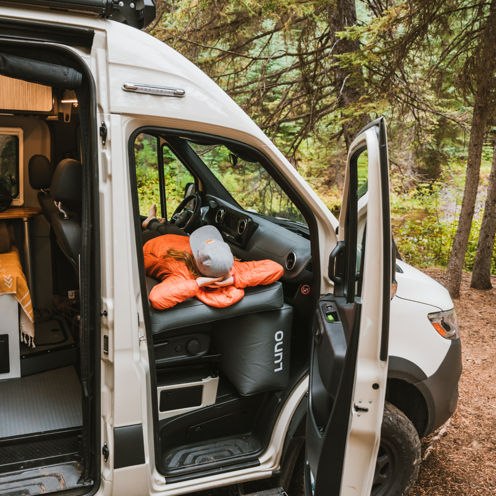 A white camper van with its front doors open, revealing an interior with bedding. A gray cap and an orange sleeping bag are placed on the front passenger seat. The van is parked in a wooded area with green trees and shrubs in the background.