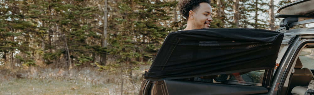 A person standing outdoors next to an open car door with a black cover partially draped over it. They appear to be preparing the vehicle. Tall trees and a forest underbrush are visible in the background.