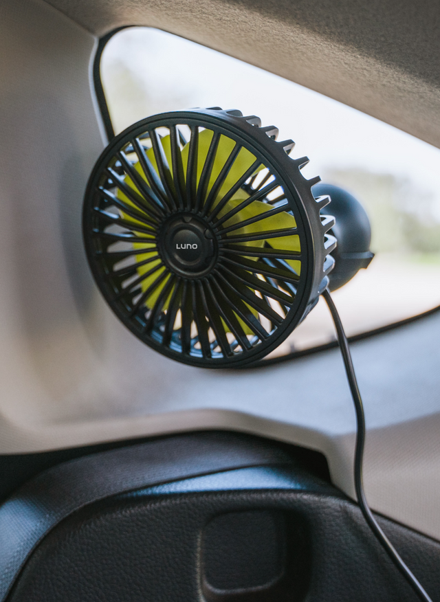 A black, portable fan mounted on the interior of a vehicle near a window, attached with a suction cup. Its power cord is visible, running down towards the base of the window. The fan's brand name "Luno" is visible at the center.