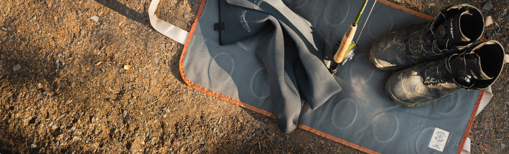 A pair of worn hiking boots, a fishing rod with a reel attached, and a piece of gray cloth are laid out on a gray mat with an orange border. The mat is placed on a dirt surface, suggesting an outdoor setting.