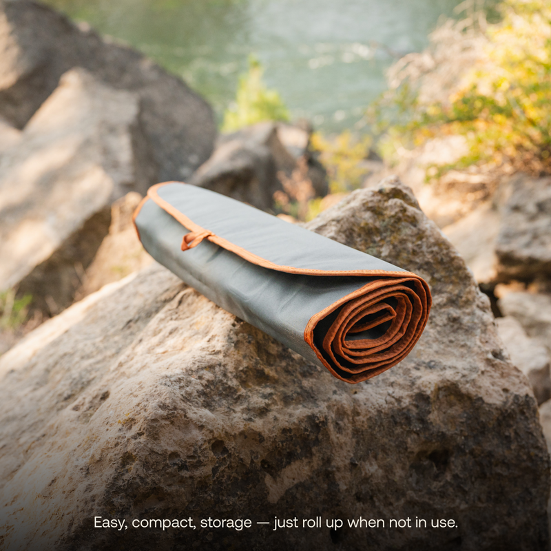 A rolled-up, compact, and durable Large Outdoor Ground Mat by Luno HQ with a gray exterior and orange trim sits on a rock near a river. The mat emphasizes easy storage and portability. Text overlay reads: "Easy, compact storage — just roll up when not in use.