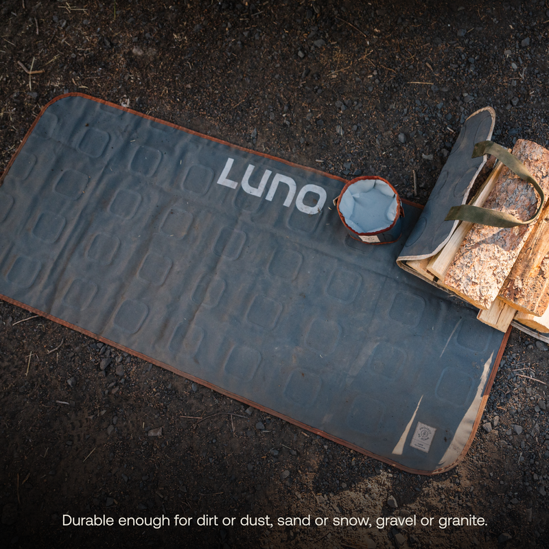 A durable multi-purpose mat made from upcycled mattresses with "Luno HQ" printed on it lies on the gravelly ground, next to a small bowl and a bundle of firewood. The caption reads: "Durable enough for dirt or dust, sand or snow, gravel or granite.
