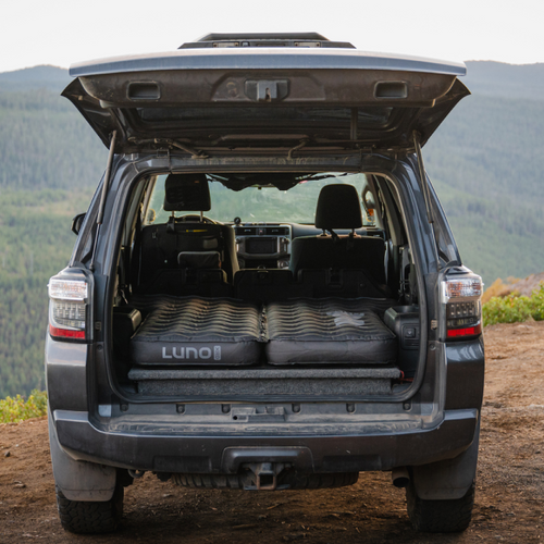 A dark-colored SUV parked outdoors with its rear hatch open, revealing a neatly laid out inflatable mattress branded "Luno" inside the cargo area. The vehicle is situated in a scenic, mountainous landscape.