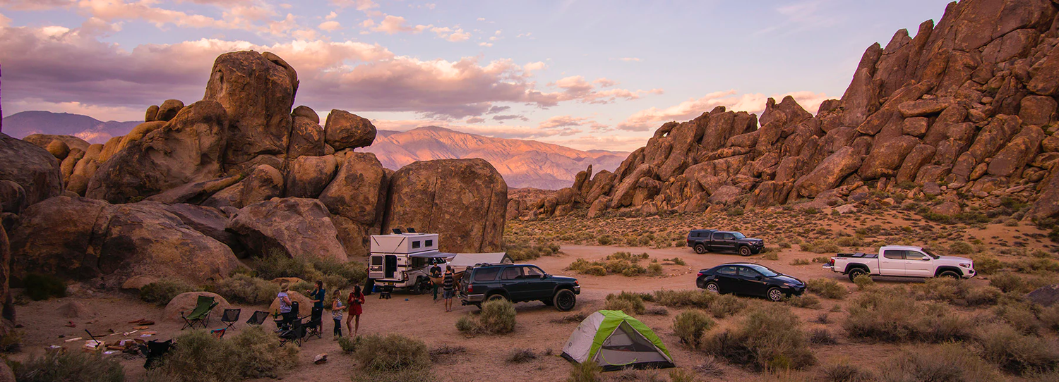 A desert campsite with several vehicles, including a camper and SUVs, surrounded by large rock formations. Tents are set up, and people are gathered near a campfire with chairs. A mountain range in the background is bathed in the warm light of the setting sun.