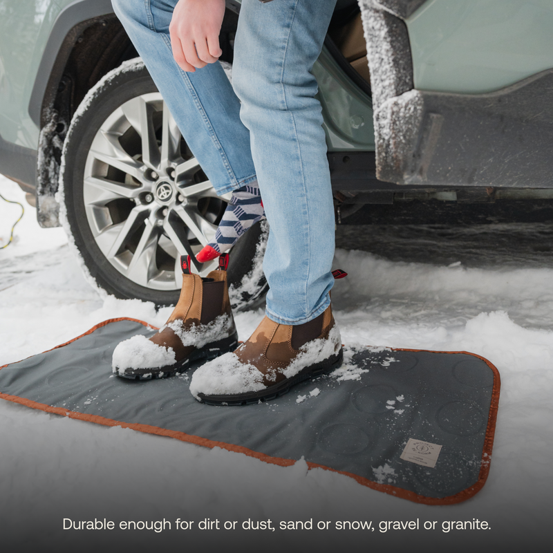 A person wearing brown boots and light blue jeans stands on a black Luno HQ Medium Outdoor Ground Mat outside a car in the snow. Snow is visible on their boots and mat. The caption reads, "Durable enough for dirt or dust, sand or snow, gravel or granite." Discover the versatility of our eco-friendly Medium Outdoor Ground Mat from Luno HQ.