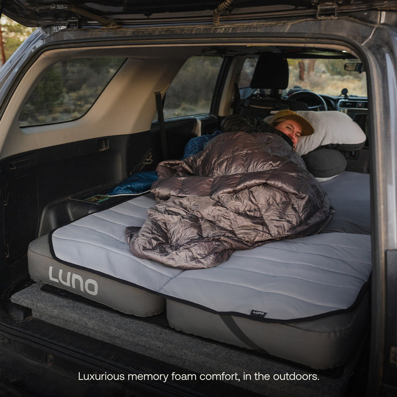 A person is comfortably lying on a Luno® Memory Foam Topper set up in the back of an SUV, bundled in a sleeping bag with a pillow under their head. The scene is cozy, set outdoors, with the tagline, “Luxurious memory foam comfort for car campers.”