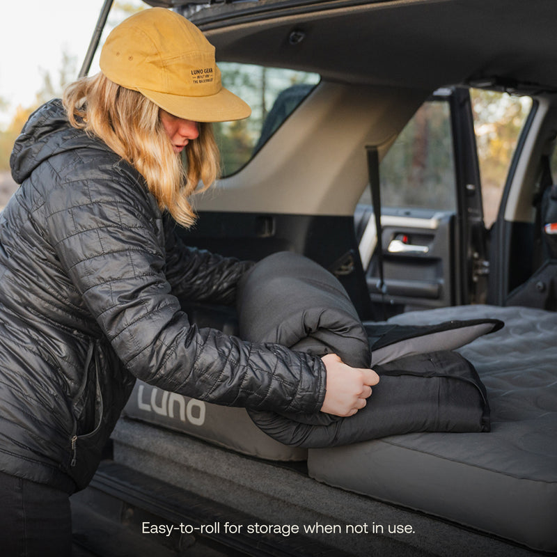 A person wearing a yellow cap and black jacket is rolling up a dark-colored Luno® Memory Foam Topper in the back of an SUV. The text at the bottom reads, "Easy-to-roll for storage when not in use." Perfect for car campers!