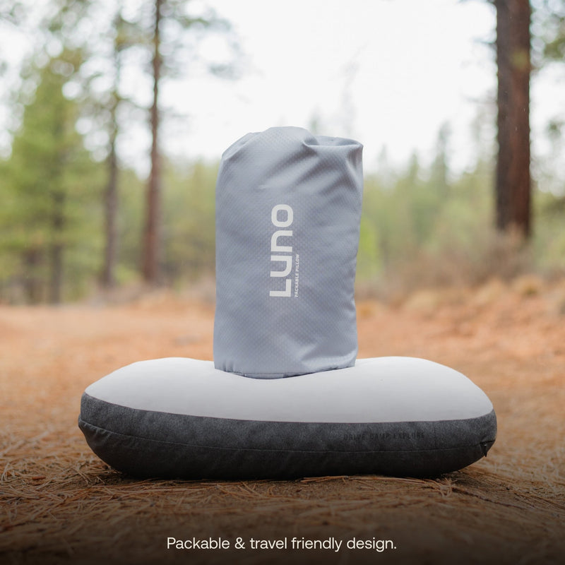 A luxury portable pillow, featuring the Luno HQ brand name and adorned in a stylish combination of gray and black, rests on a forest floor amidst the trees. On top of it is a gray cylindrical bag. Text reads, "Packable & travel-friendly design with quick USA shipping.