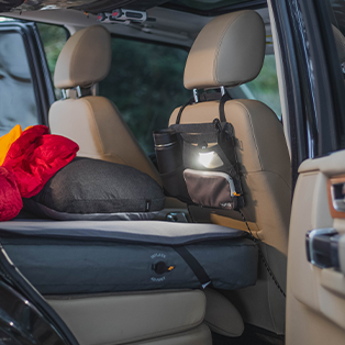 The back seats of a car with the rear door open, showing a setup for car camping. An inflatable mattress covers the back seat, with a red sleeping bag and black pillow on it. A small organizer is attached to the back of the front passenger seat, holding a flashlight and drink bottle.