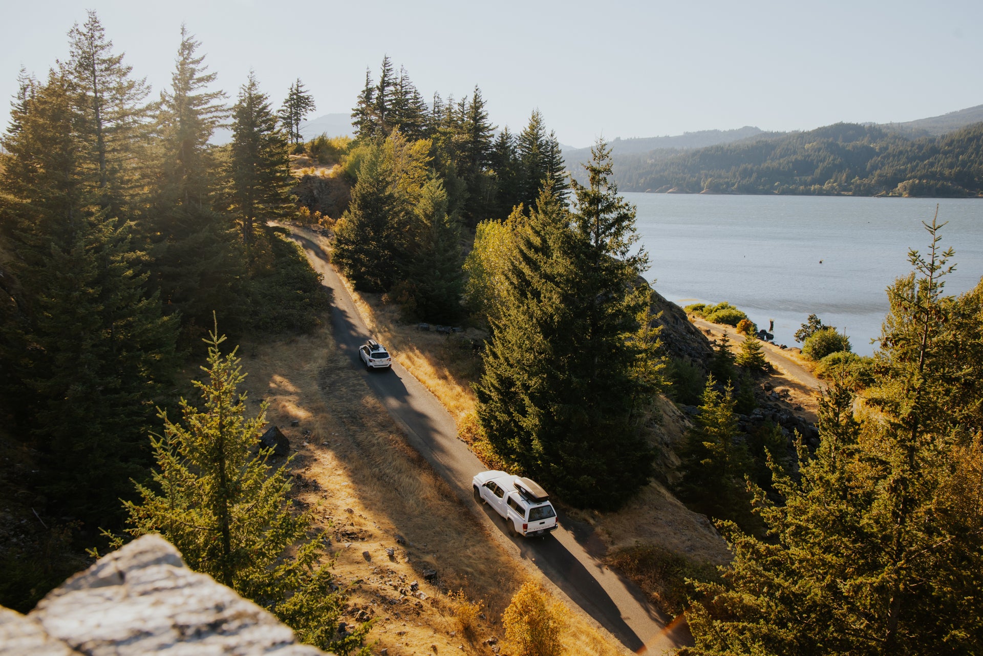 A scenic landscape depicting two cars driving along a narrow, winding road surrounded by tall evergreen trees. The road overlooks a large body of water, with hills visible in the background under a clear sky.