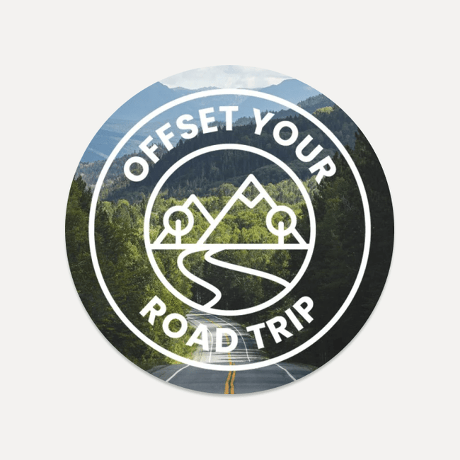 A circular logo depicting a winding road through a lush, mountainous landscape features the text "OFFSET YOUR ROAD TRIP" encircling a stylized image of mountains and trees with a road winding through them, all bordered by white. This design accentuates the journey toward being Carbon Positive with Road Trip Carbon Credits from Luno®.