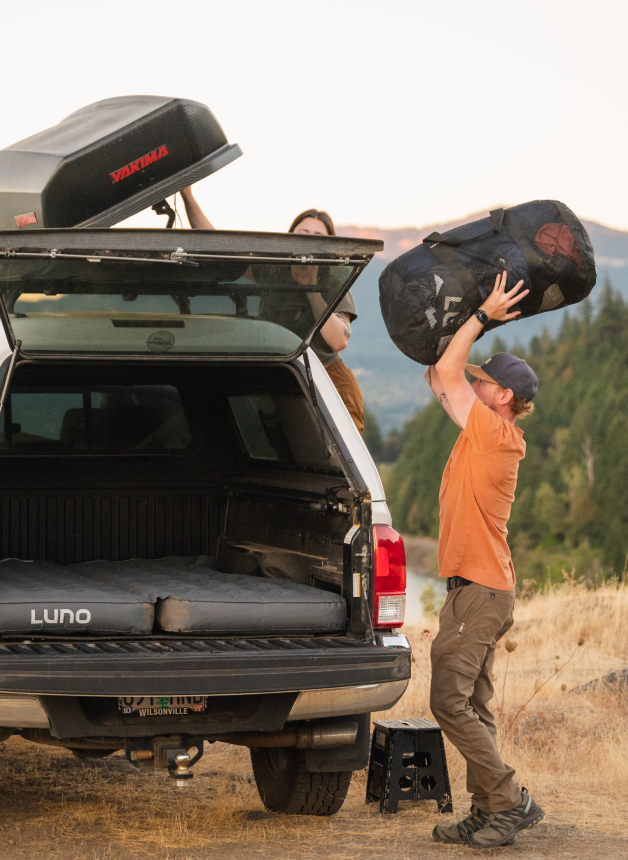 Two individuals are packing a rooftop cargo box on top of a white Luno-equipped pickup truck parked in an outdoor setting. One person is lifting a large duffel bag towards the other who is on the truck bed. They are surrounded by dry grass and trees.