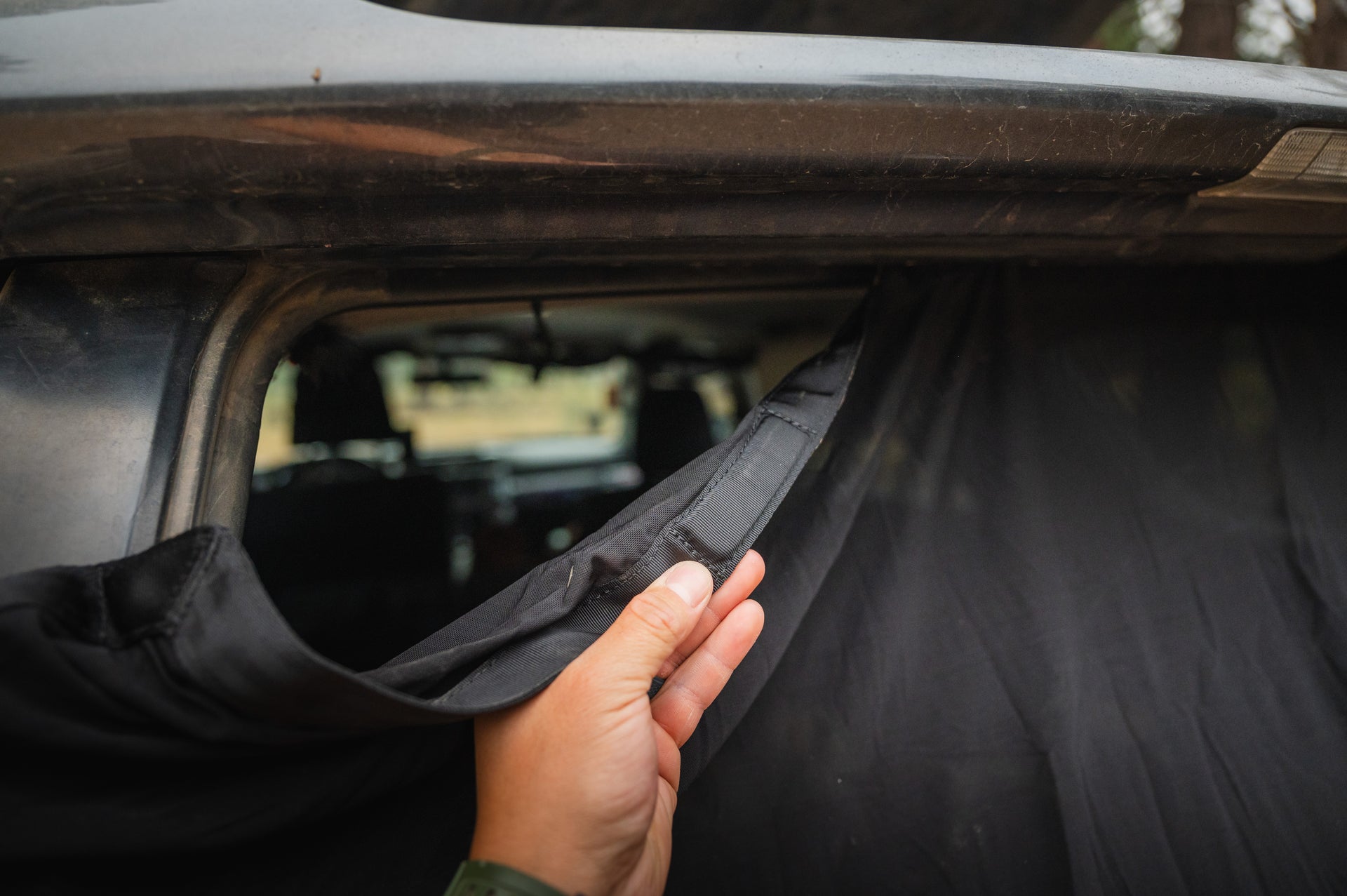 A close-up of a person lifting a black curtain or cover attached to the inside of a dusty car's rear window. The interior of the car is dimly lit and partially visible through the uncovered portion of the window.