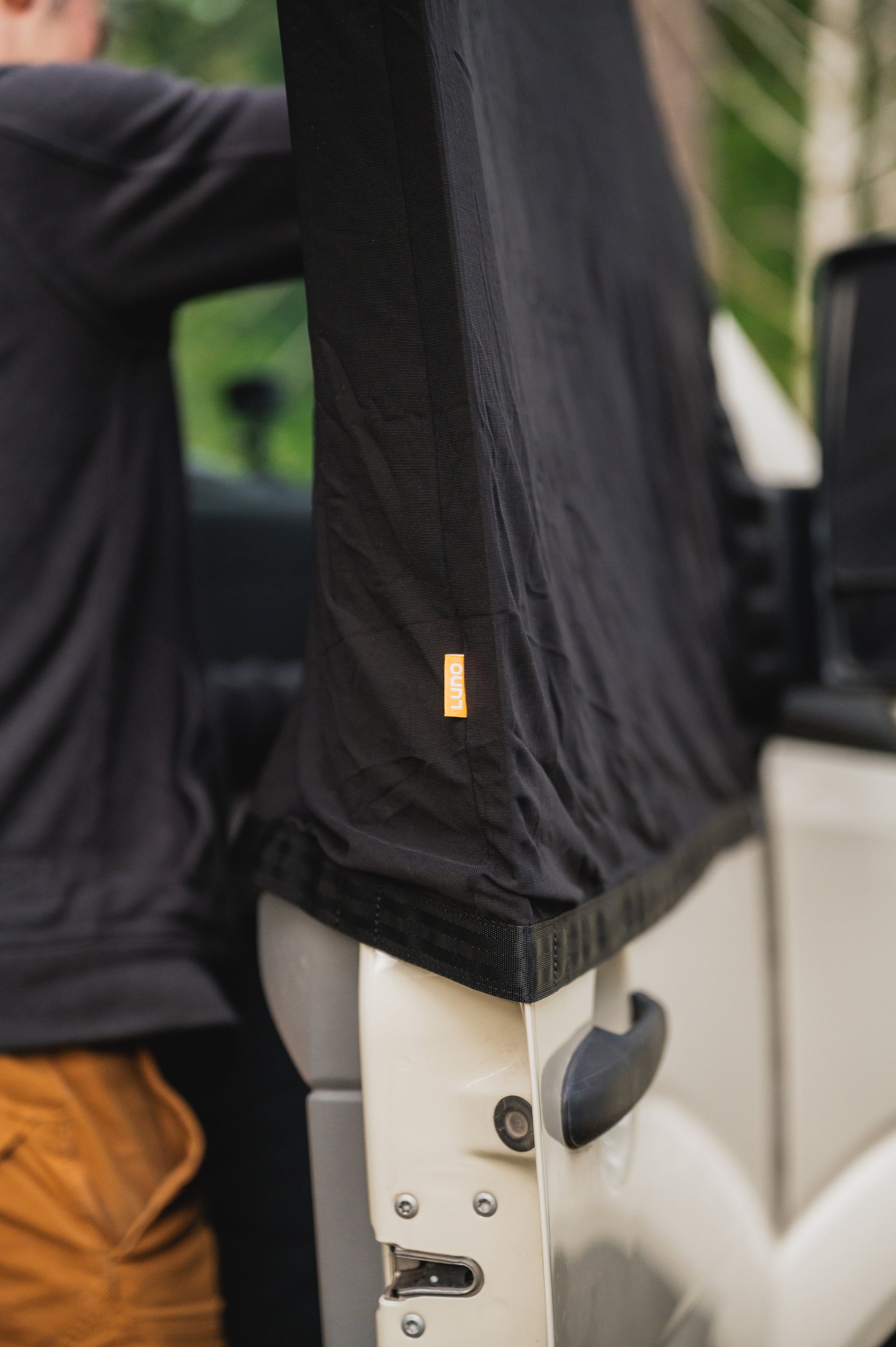 A person is setting up a black fabric cover over the top portion of a white car door. The cover has an orange tag with text on it. The person is wearing a black long-sleeve shirt and brown pants. The background includes some green foliage.