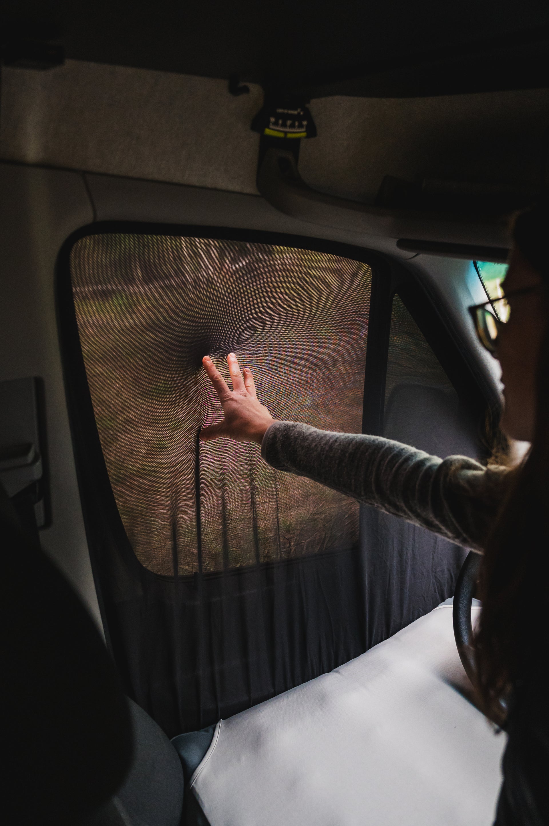 A person wearing glasses and a striped shirt is pressing their hand against the mesh of a car window sunshade from inside a vehicle, creating a ripple effect on the mesh. The interior of the vehicle is partially visible, including the dashboard and steering wheel.