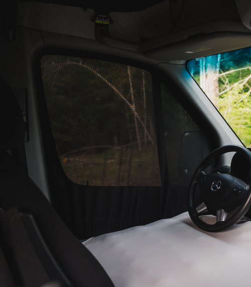 Interior of a vehicle, focusing on the driver's seat with a black steering wheel and a mesh screen covering the side window. The scene outside is blurred, with hints of green foliage and wooden branches visible through the mesh.