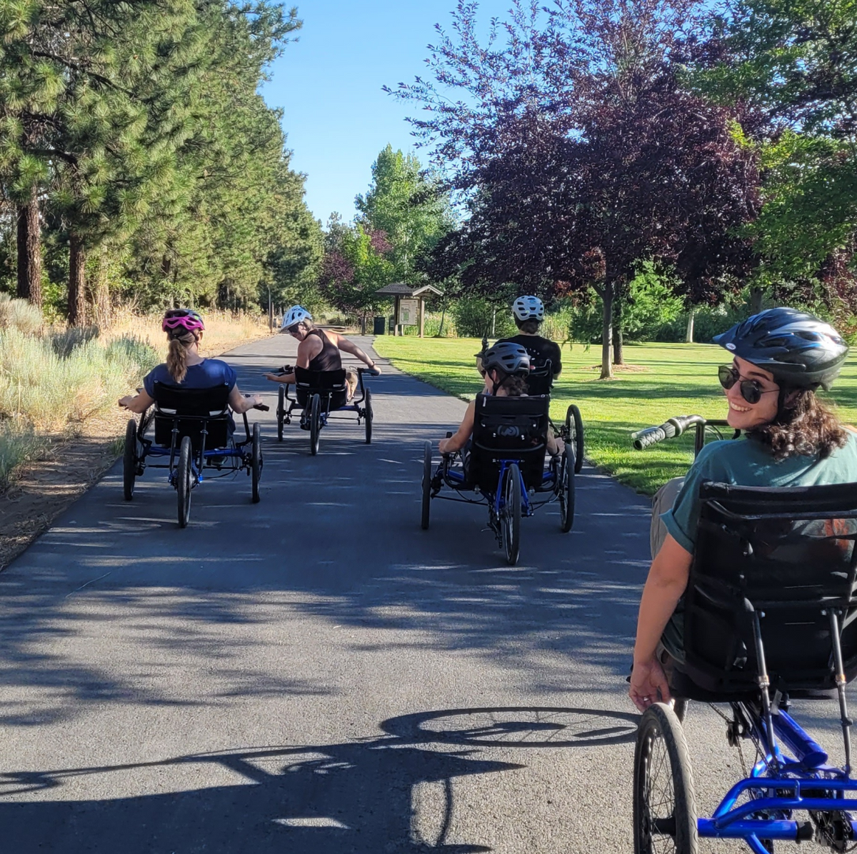 A group of people riding recumbent trikes on a paved path in a park. They are wearing helmets and are surrounded by trees and greenery. The person in the front right is looking back and smiling at the camera.