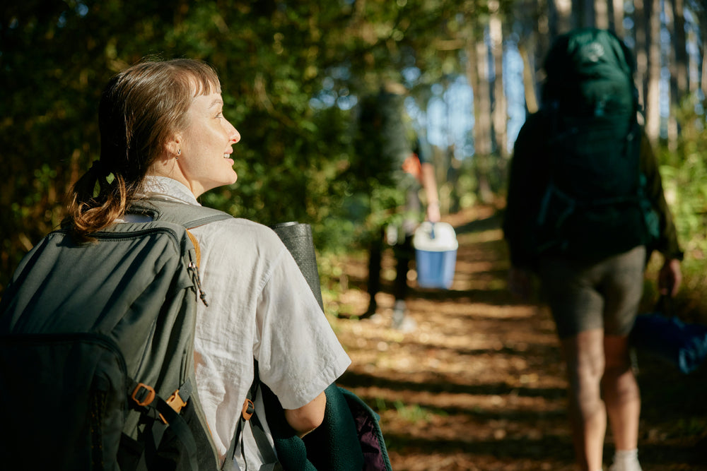 A person with a backpack is walking on a forested path, smiling and looking to the left. Another person is ahead, carrying a cooler. The trail is surrounded by tall trees, and sunlight filters through the branches, creating a serene outdoor hiking scene.