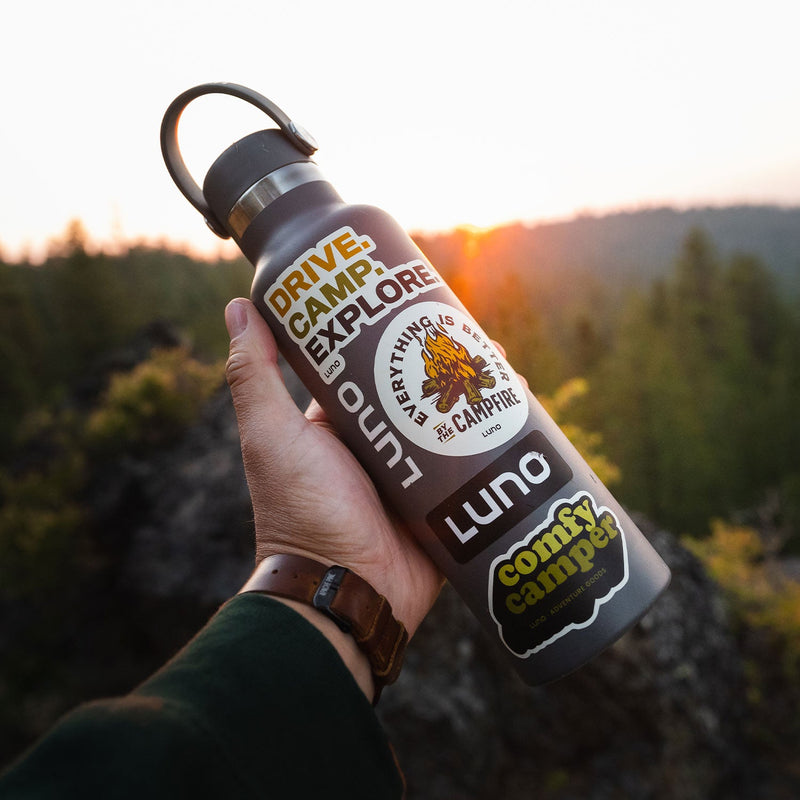 A hand holds a gray water bottle decorated with Luno HQ's Camping Sticker Pack—featuring camping-themed designs—set against a blurred forest and bright sunset, perfect for any car camping adventure.